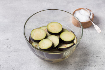 Soaking eggplant slices in salt water in a glass bowl on a gray textured background. Processing eggplant before cooking, removing bitterness