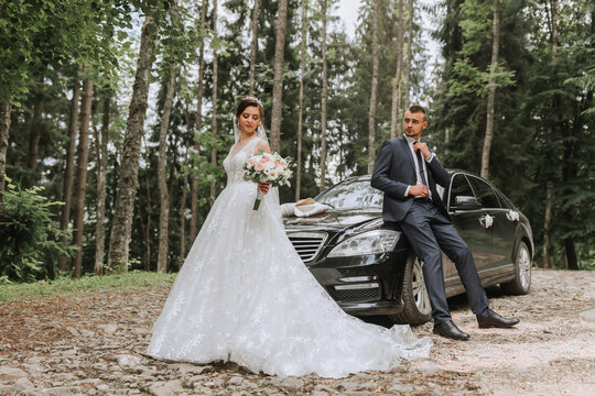 Front view of a married bride and groom wearing festive clothes standing against a black car on their wedding day