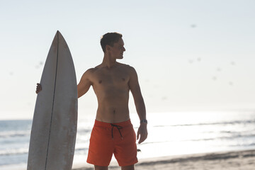 A shirtless man surfer standing on the beach holding a surfboard - looking to the side - Powered by Adobe