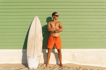 A smiling man with nice body wearing sunglasses standing at the light green with a surfboard - looking to the side - Powered by Adobe