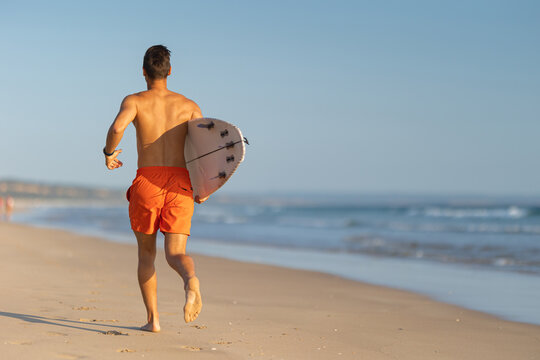A man with nice athletic body running on the shore holding a surfboard