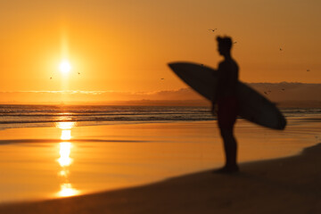 Black silhouette of a man standing on the shore holding a surfboard at orange sunset