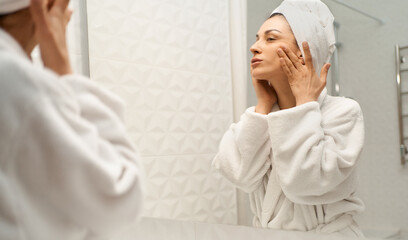Adult woman wrapped in white bath towel in bathrobe in front of mirror lifting and massaging her skin. Concept rejuvenation and skin lifting