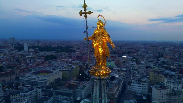 Aerial view of the statue of the Madonna on the central spire of the Duomo and the city's cathedral at dawn. Roofs of houses and skyscrapers. Sun over the horizon. duomo square. Italy Milan 05.08.2023