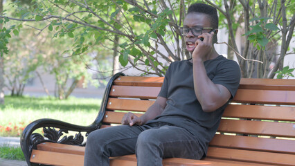 Obraz premium Upset Young African Man Arguing on phone while Sitting Outdoor on a Bench