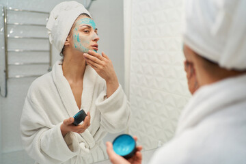 Adult lady stands in front of mirror gently applying a natural face mask on facial skin. Beauty of home skincare rituals and feel rejuvenated
