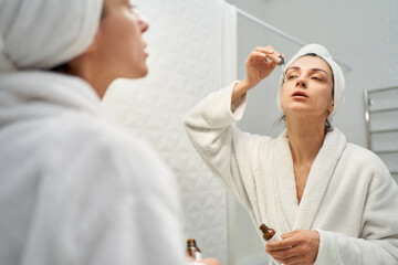 Stunning woman in bathrobe applying serum to her face as part of her daily skincare routine. Perfect for beauty and skincare related projects