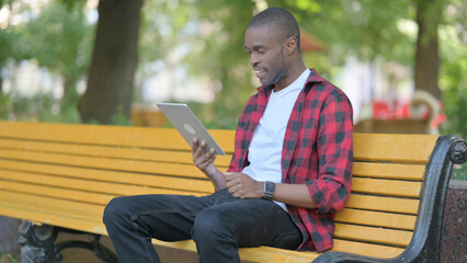 Obraz premium Young African Man Doing Video Chat on Tablet while Sitting Outdoor on a Bench