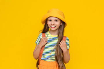 A little girl scout, smiling happily in a summer hat with a backpack. Hiking for children in nature. Yellow isolated bright background.