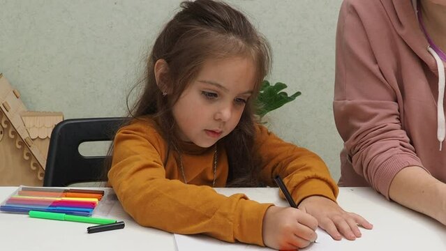 Small preschooler girl sit at desk study online on phone with mom, smart little kid  learns  internet lessons on quarantine, homeschooling. Draws with felt-tip pens and colored pencils, imagination