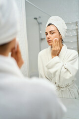 Multiethnic woman wearing towel on head using cotton pad for facial skin care and looking in mirror in bathroom - 631530515