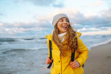 Young woman tourist in a yellow coat walks along the seashore, enjoys the seascape at sunset. Travel, tourism concept. Active lifestyle. - obrazy, fototapety, plakaty