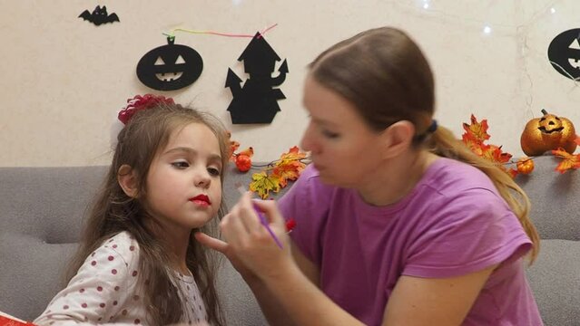 Mother making Halloween makeup to preteen girl daughter and painting spooky black lines on face. Pretty child with creepy cosmetics preparing for October holiday party