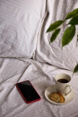 Smartphone and a cup of coffee placed on a bed with white sheets in the background