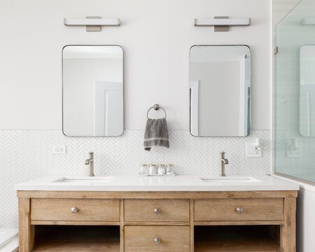 A cozy bathroom detail with a white oak vanity cabinet, white countertop, bronze faucets and lights and a white herringbone tile backsplash.