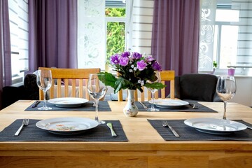 Dining table with plates, wine glasses, and a bouquet of purple flowers matching with curtains.