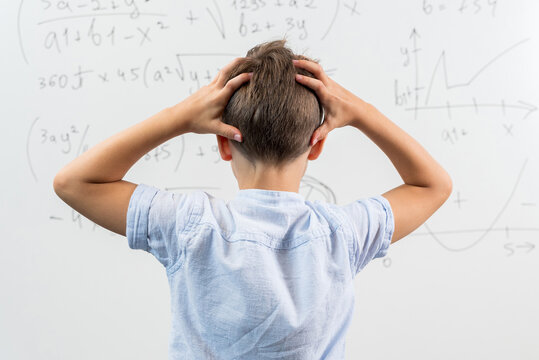 View from the back of schoolboy standing in class against blackboard. Boy holding hands on head. Portrait of shocked and emotional little kid. Back to school. Failing to solve a mathematical equation