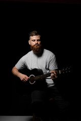 Bearded brutal guitarist plays an acoustic guitar in a black room.