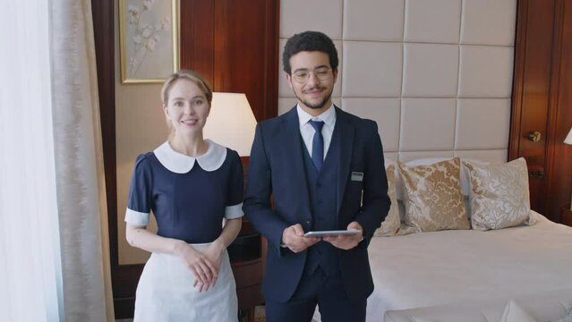 Portrait of cheerful hotel manager and smiling maid standing together in room and looking at camera, bedroom interior in background, man holding digital tabletPortrait of cheerful hotel manager and sm