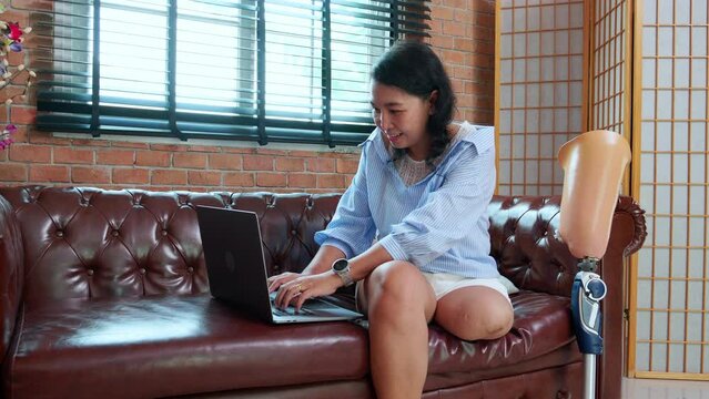 Young asian woman disabled sitting on sofa using laptop computer with prosthetic leg in living room at home, female working on notebook and prosthesis with optimistic, lifestyles and disability.