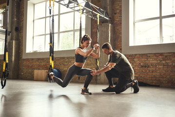 Doing squat exercise. Confident young personal trainer is showing slim athletic woman how to do...