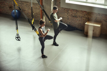 Obraz premium Exercising together. Young active couple in sportswear training legs with Trx fitness straps at gym.