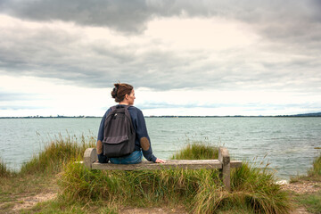 Lone woman sitting on a bench by the water, enjoying the view. - obrazy, fototapety, plakaty
