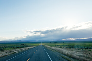 A long asphalted straight road stretches far into the horizon, on which blue mountains can be seen. The rays of the sun break through the large clouds over the road. blue sky before sun