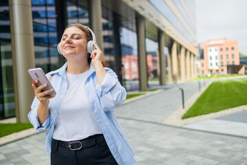 Fototapeta premium Young happy woman wearing suit using wireless earphones holding smartphone listening to music and singing, walking on urban city street. High quality photo