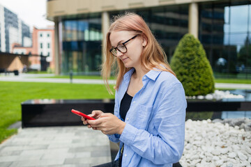 Cheerful happy stylish woman using smartphone online app while sitting near the mall. Happy woman in a blue shirt walks down the street looking at her smartphone. An exciting application used for