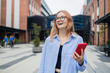 Cheerful happy stylish woman using smartphone online app while standing near the mall. Happy woman in a blue shirt walks down the street looking at her smartphone.An exciting application used for