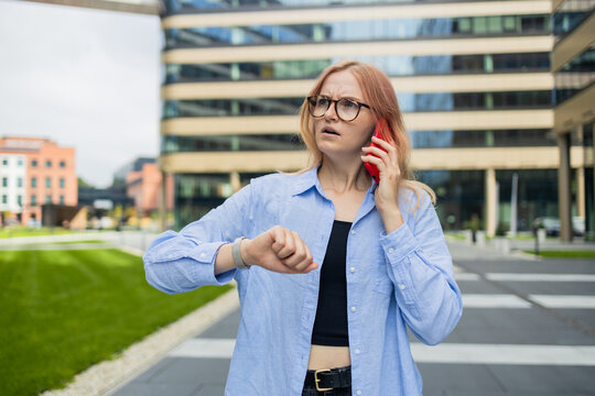 Angry irritated young business woman holding mobile phone and looks at his watch at the blurry business center building. High quality photo