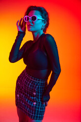 Portrait of woman wearing fashionable touching sunglasses in transparent top and skirt posing over gradient yellow-red neon background.