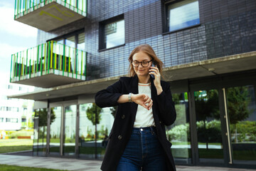 Fototapeta premium Smiling blonde business woman hurry up to airport, standing on city street with phone in hand, looking at time on hand watch while calling taxi by mobile phone. Business trip concept
