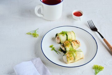 Fried chicken rolls wrapped with sliced zucchini on a white plate on a light concrete background. Recipes for zucchini.