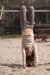 Girl child hanging upside down on a bar in the playground touching the sand with her hands in...