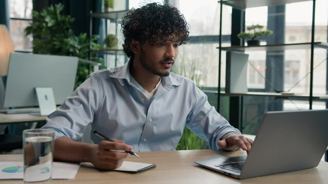 Arabian Indian latin man businessman guy write business notes ideas in notebook managing corporate tasks writing schedule with pen on desk male worker employee working with laptop computer in office