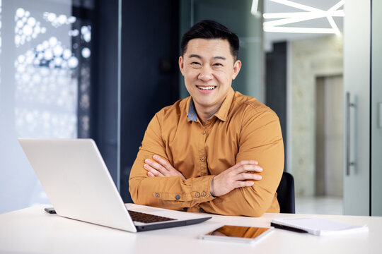 Portrait of mature experienced Asian businessman inside office at workplace, boss programmer sitting at desk with arms crossed smiling and looking at camera with laptop.