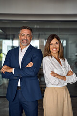 Vertical portrait of smiling mature Latin hispanic business man and European business woman standing arms crossed in office. Diverse colleagues, group team of confident professional business people.
