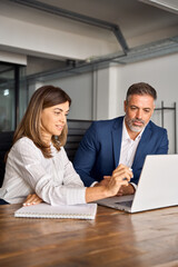 Obraz premium Vertical shot of colleagues, group of professional business people working together. Mid age European business woman mentoring mature 50s age Latin business man discussing project on laptop in office.