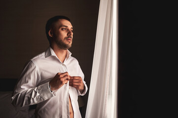 Handsome young man groom button up white shirt in hotel room, serious looking at window. Confident...