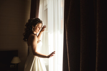 Portrait of woman bride in wedding dress with nice makeup and hairstyle at window, pensive closed...