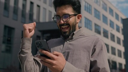 Indian Arabian man guy employer entrepreneur shock amazing read news on mobile phone winning online bet business achievement good job offer happy excited businessman in city outdoors celebrate victory