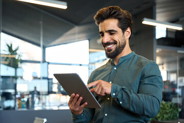 Happy young Latin business man executive holding pad computer at work. Male professional employee using digital tablet fintech device standing in office checking financial online market data. - obrazy, fototapety, plakaty