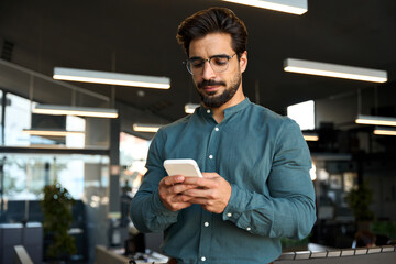 Busy young Latin business man using cellphone at work standing in office. Serious male executive,...