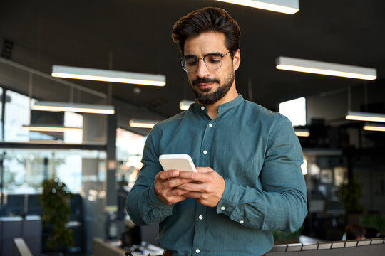 Busy young Latin business man using cellphone at work standing in office. Serious male executive, businessman employee or entrepreneur holding smartphone working on mobile cell phone at work.