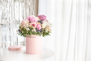 An elegant bouquet of fresh roses, peonies, and daisies in a pink box, beautifully arranged and placed by a light-filled window.