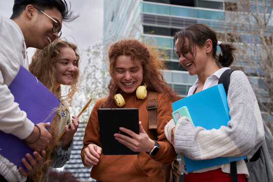 Group Of Cheerful Students Sharing An Digital Tablet To Check Internet. They Are Standing Up Outside The Campus. They Check Social Media And All Look Happy