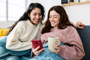 Happy young female friends using mobile phone sitting together on sofa at home. Winter domestic...