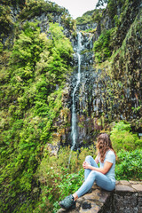 Female tourist sits on stone wall viewpoint and watches green overgrown Risco waterfall. 25 Fontes...